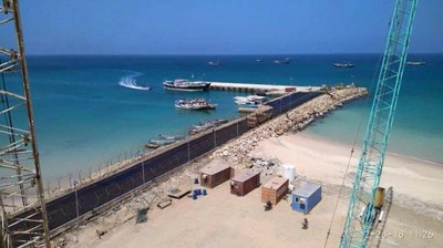 A long breakwater with a dock at the end and commercial boats offloading ,in theforeground a beach area with containers