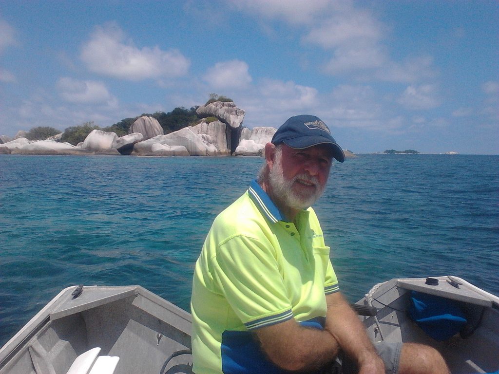 a man sitting in his dinghy in front of a group of boulders with a white sandy beach