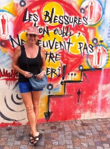 lady in shorts, sleeveless top and pink cap stands in front of a wall covered in brightly covered graffiti