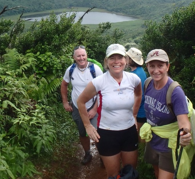 A group of 4 people hiking up a green lush mountainside with a view of a lake below, happy and smiling