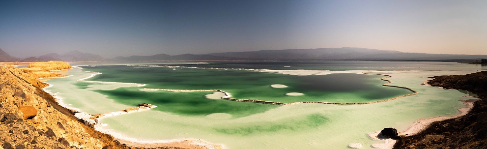 Green blue water surrounded by yellow sand and arid rock with white crusts of salt around its edges