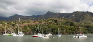 green shoreline with thick forest rising to a dark cloudy peak behind and in the foreground still waters with several boats at anchor