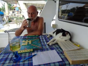 man with no shirt on and a bald head with white beard sitting at a table with a blue checked tablecloth on the aft deck of a boat. He is drinking a cup of coffee and a cat is lying on the table. Looks like scrabble is being played