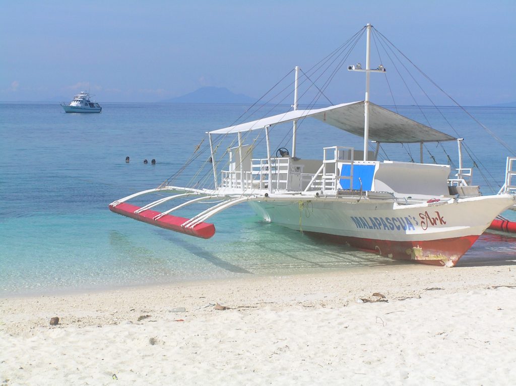 white powder beach with a local boat with outrigger pulled up on it and a motorboat at anchor in the distance across a blue blue sea