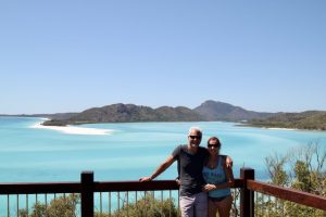 a couple with their arms around each other standing in front of a wooden fence with pale turqoise sea behind them and a low mountainous earthy coloured island