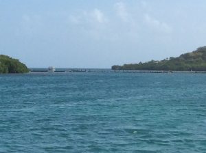 an expanse of shallow bright blue water with 2 islands in the distance connected by a wobbly looking wooden footbridge